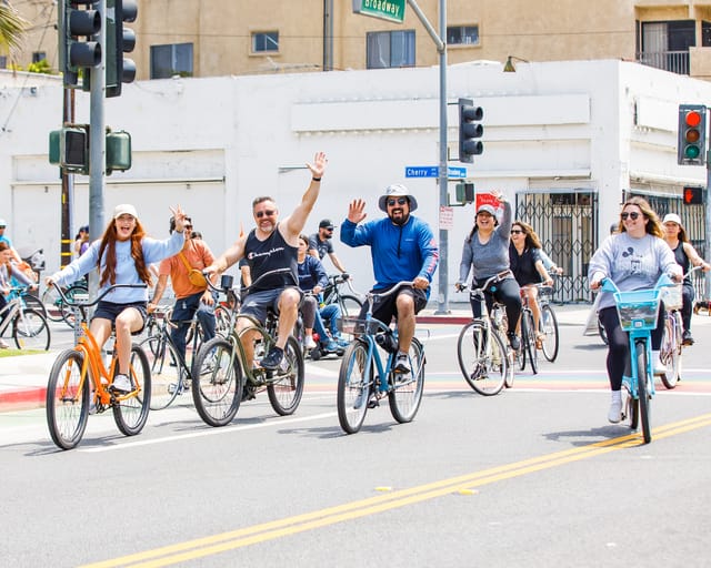 People, many smiling and waving, riding bicycles down the street.