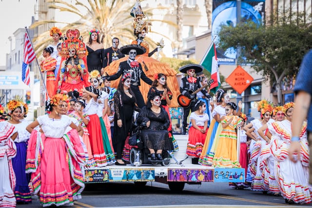 People in colorful garb and makeup ride a float down a downtown street.