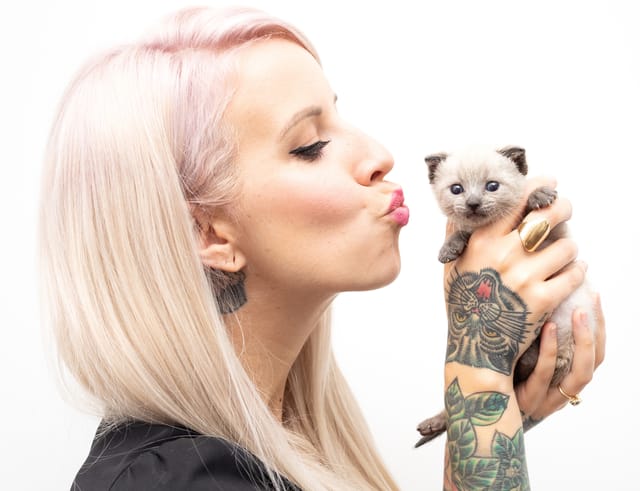 A woman holds up a tiny gray kitten as if to kiss it.