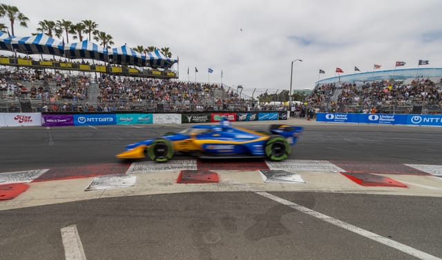 A blue and orange race car speeds past grand stands.