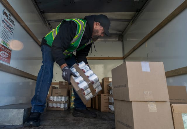 A bearded man wearing a black hat moves a box of canned food in the back of a moving truck.