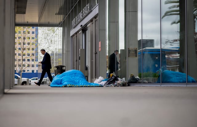 A man in a suit walks past a person sleeping under a blue blanket outside a glassy office building.