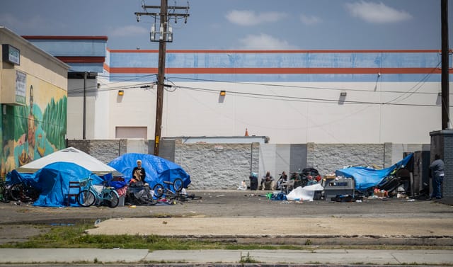 People sitting near blue tents and other items on a vacant city lot.