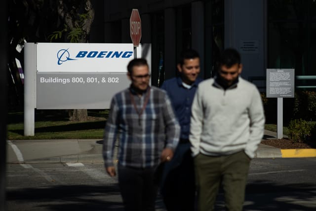 Three out of focus people walk away from a white and blue sign that reads "Boeing."