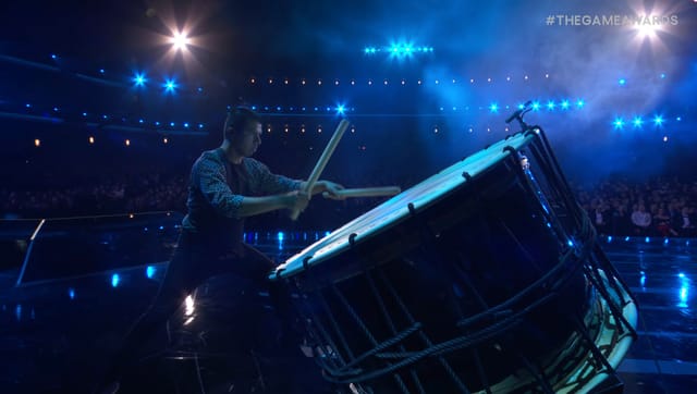 A man plays a large drum with white and blue lights shining down on him.