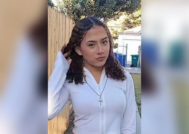 A teen girl wearing a white long sleeve shirt standing outside next to a fence.