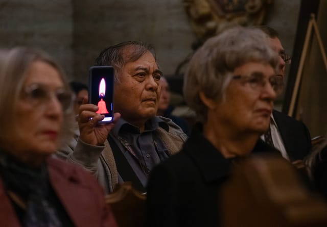 A man standing in a crowd holds up a phone displaying the image of burning candle.