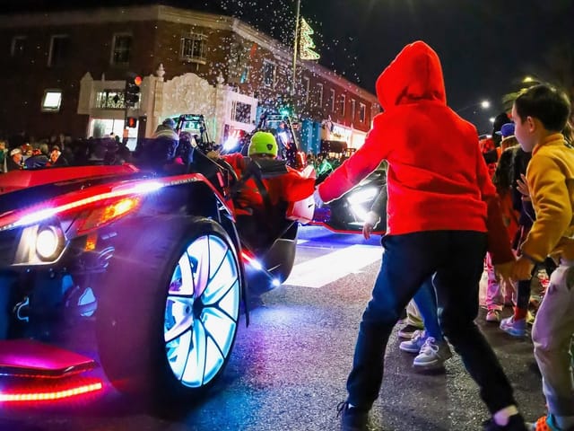 People get up close to a brightly lit car during a nighttime parade.