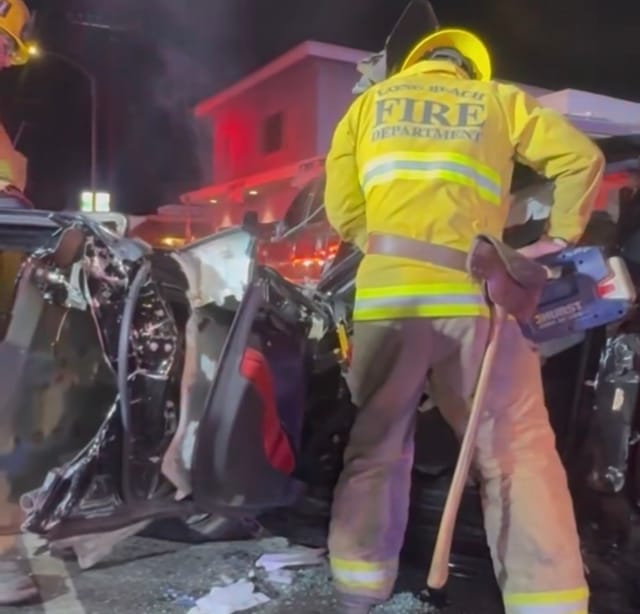 A person in a yellow jacket that says "Long Beach Fire Department" on the back stands next to a wrecked car.