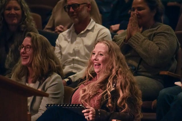 A red-haired woman smiles broadly while sitting surround by other people.