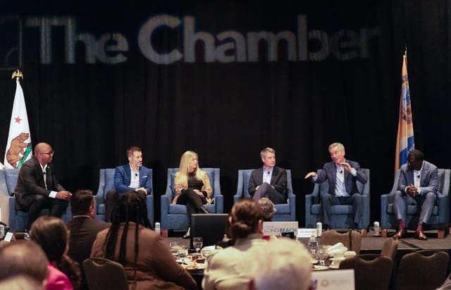 Six people in business clothes sit on a stage in front of a large audience.