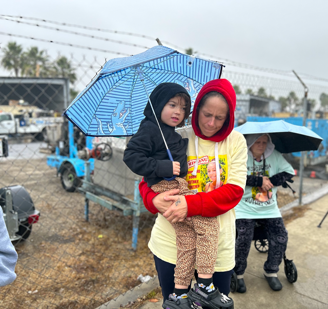 A woman inna red hoodie holds a blue umbrella and a small child in a black hoodie.