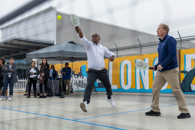 Two men hold white paddles and play a game a pickleball in business casual clothes.