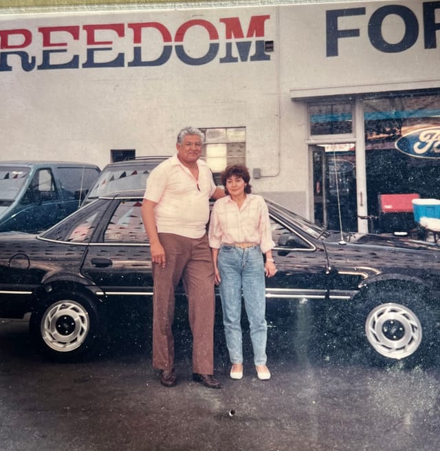 A man and a woman stand in front of a black car.