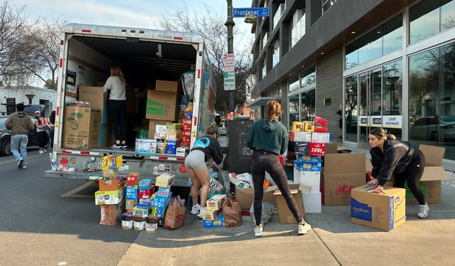 People loading stacks of boxes of food on the sidewalk oto a delivery van also carrying boxes.