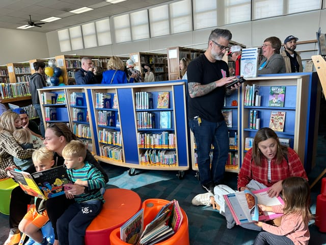 Children and adults sit on colorful round benches near many shelves of books.