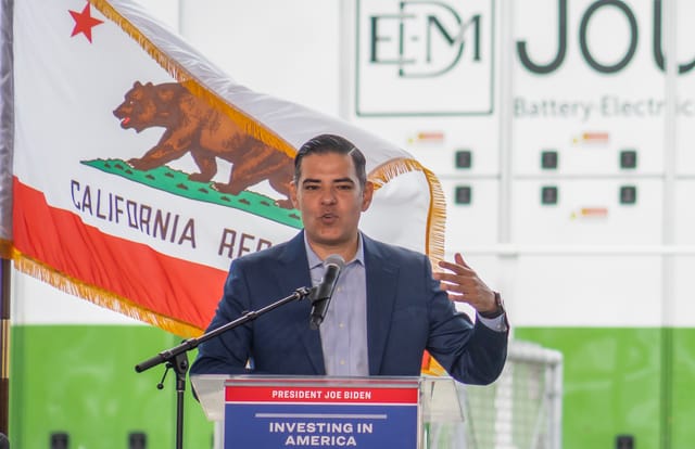 A man speaks at a microphone with the California flag waving behind him.