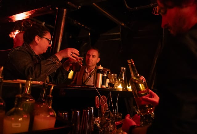 A man holds a bottle while two people clink glasses in a dark room.