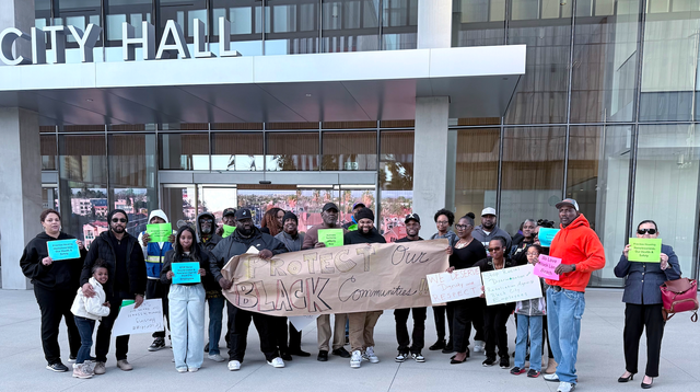 A couple dozen people holding signs stand in front of a glassy office building labeled "City Hall.