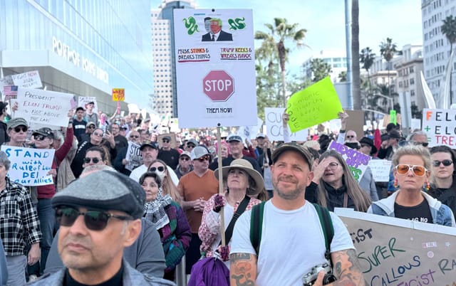 Hundreds of people stand outside and hold political signs near an office building.