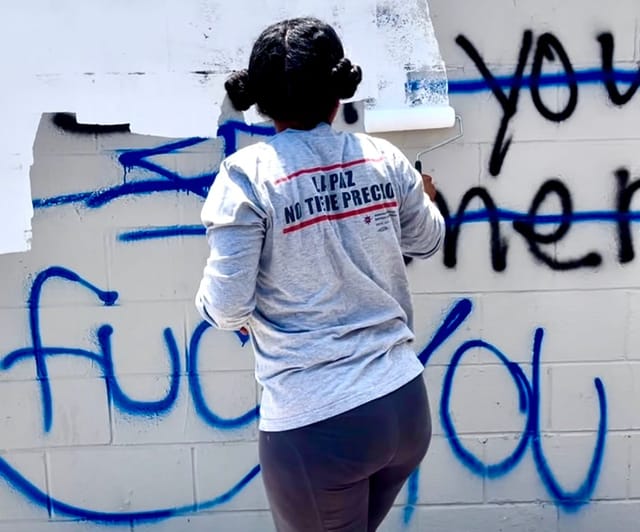 A woman paints over graffiti on a white brick wall.