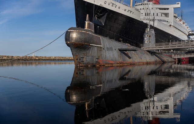 An old, rusty submarine sits beside a historic black and white ocean liner with the name "Queen Mary" written on it.