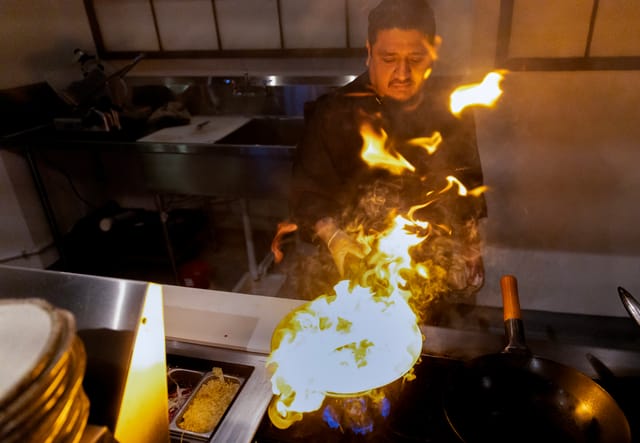 A man stands next to a stovetop with a pan that is on fire.