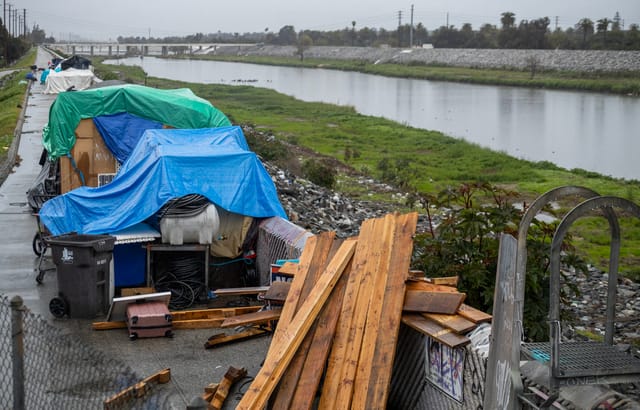 A homeless encampment with makeshift structures covered in blue and green tarps sits beside a river.