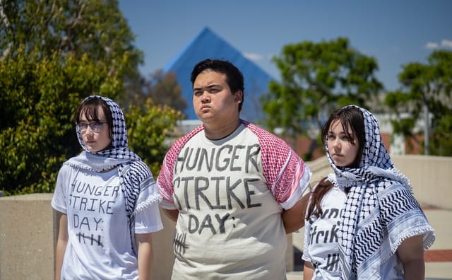 Three young adults stand shoulder to shoulder each wearing a t-shirt that says "hunger strike day."