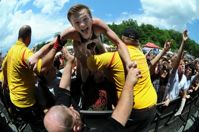 A smiling person is crowd-surfing and being propped up by two men in yellow shirts.