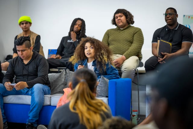 A group of young people sit thoughtfully around one person speaking into a microphone.