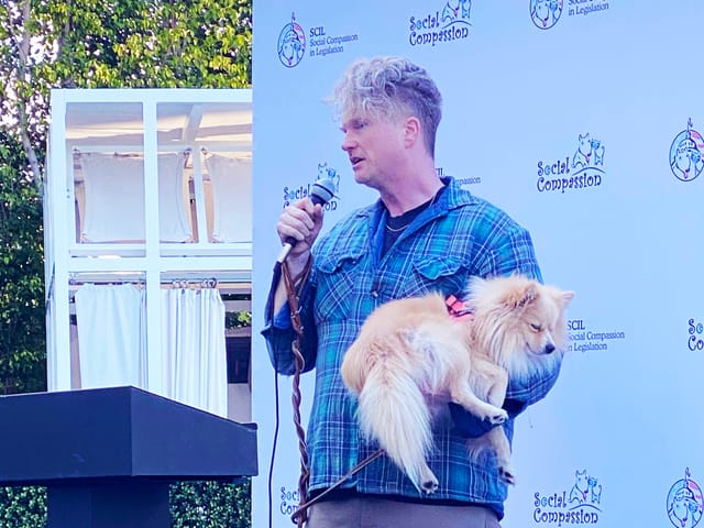 A man stands at a lectern holding a microphone and a small beige dog.