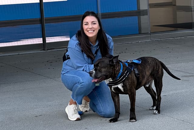 A woman is kneeling down next to a really sweet black dog.