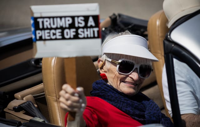 A woman wearing sunglasses and a white visor sits in a convertible car while holding a sign saying "Trump is a piece of shit."