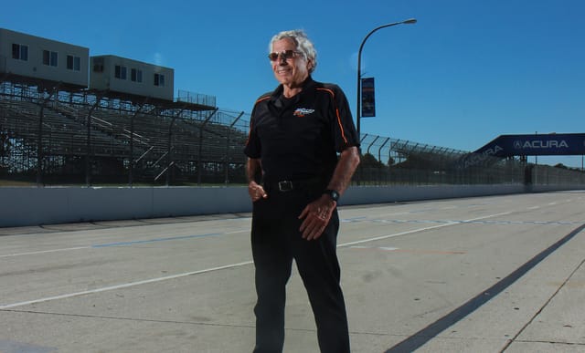 A gray-haired man smiles as he stands alone on an empty race track on a sunny day.